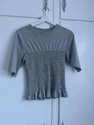 Grå ribbad topp med volangkant - Säljer en grå topp med ribbad struktur och smockad midja. Toppen har korta ärmar, rund halsringning och en söt volangkant nedtill. Perfekt för dig som gillar en enkel men ändå detaljrik stil.