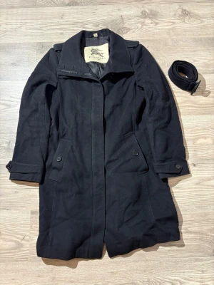 Burberry  - Stilren svart kappa från Burberry i ullmix med klassisk krage, knappar vid ärmslut och fickor samt tillhörande bälte. Perfekt för dig som vill ha en tidlös och elegant look. Kappan har lång ärm och en rak passform.