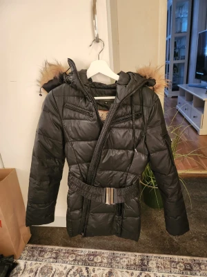 Svart dunjacka med fuskpäls och bälte - Snygg svart dunjacka från Only med avtagbar huva och fuskpälsdetalj. Jackan har quiltad design, dragkedjor på bröstet och midjebälte för en schysst passform. Perfekt för kalla dagar och riktigt varm.