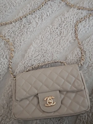 Beige quiltad handväska - Supersnygg beige quiltad handväska med klassisk kedjerem i guld och ikonisk CC-logga framtill. Väskan har lock och elegant rutmönster, perfekt för dig som vill ha en tidlös och lyxig accessoar. Materialet är mjukt skinn som känns exklusivt.