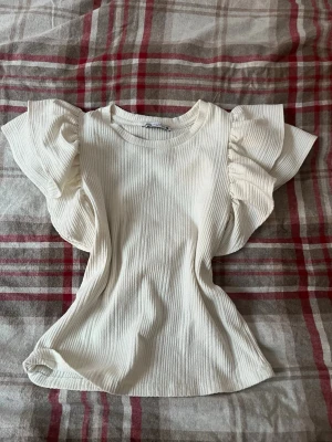 Vit ribbad topp med volangärmar💓 - Superfin vit ribbad topp med stora volangärmar. Toppen har rund halsringning och är tillverkad i ett mjukt material som ger en bekväm passform. 💗💗 Den har en liten fläck som jag försöker visa i sista bilden, men förmodligen går o tvättas bort o e nästan inte synlig❣️