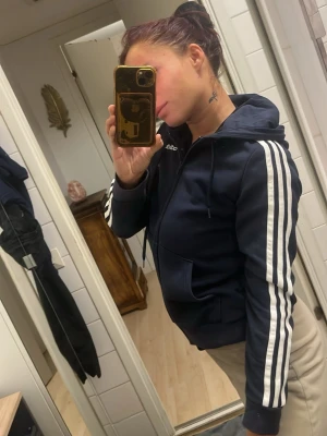 Mörkblå Adidas hoodie med dragkedja - Snygg mörkblå hoodie från Adidas med dragkedja framtill och klassiska vita ränder längs ärmarna. Huva med dragsko och två fickor framtill. Perfekt för en sportig och avslappnad stil.