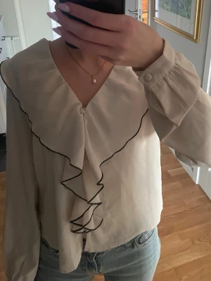 Blus med volanger  - Säljer denna söta blusen med volanger ifrån Only! Blusen är i storlek XS och använd ett få tal gånger! Säljer då den inte kommer till användning! Färgen är ljusbeige/krämvit men svarta detaljer. Blusen har även snygga klädda knappar på ärmen samt öppningen. 