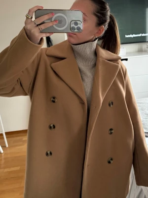 Beige kappa med dubbelknäppning - Stilren beige kappa med dubbelknäppning och bred krage. Klassisk rak modell som ger en snygg och tidlös look. Perfekt att bära över stickade tröjor under kyliga dagar. Kappan har långa ärmar och stora knappar framtill.