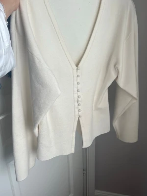 Krämvit kofta med knappar - Stilren krämvit kofta med djup V-ringning och dekorativa tygklädda knappar framtill. Koftan har långa ärmar och en mjuk, något tjockare stickad känsla. Passar perfekt till lager på lager och ger en clean look.