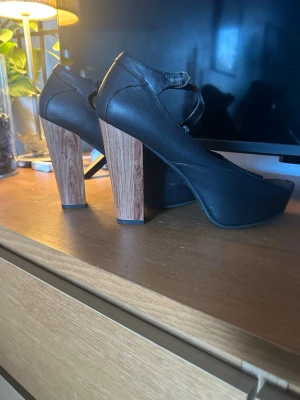 Svarta pumps med träklack - Snygga svarta pumps med bred träklack och rund tå. Skorna har en rem över foten med spänne. Skorna är använda så sulan är inte nya. 