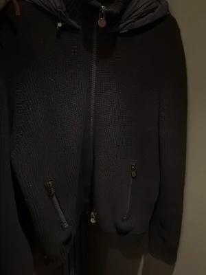 Svart stickad jacka från Moncler - Svart stickad jacka från Moncler med huva och dragkedja framtill. Jackan har två snedställda fickor med dragkedjor och Moncler-logga på ärmen. Kombinerar stickade partier med vadderad huva för en cool look.