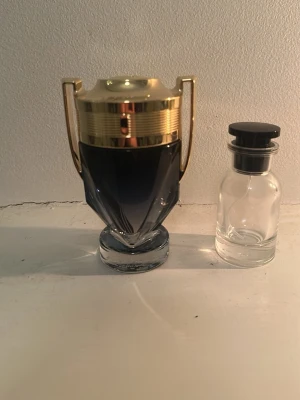  Rabanne Invictus parfymflaska - Snygg parfymflaska från Rabanne Invictus med unik troféform. Flaskan är gjord i glas med en mörkblå toning och har guldfärgade detaljer på locket och handtagen. Perfekt för dig som gillar exklusiv design på din parfymhylla.
