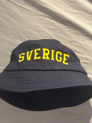 Mörkblå bucket hat med SVERIGE-tryck - Snygg mörkblå bucket hat med gul text där det står SVERIGE framtill. Klassisk form med bred skärm och sydda detaljer. Tillverkad i bomullsmix och passar de flesta huvuden tack vare one size-storlek. Perfekt för dig som vill visa ditt stöd för Sverige på ett stilrent sätt.