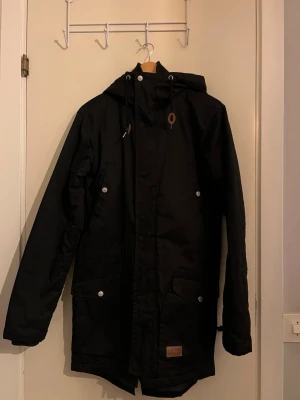 Svart parkas från Etirel med luva - Svart parkasjacka från Etirel Heritage Collection med huva och snörning. Jackan har långa ärmar, tryckknappar, dragkedja och två stora fickor framtill. Quiltad insida för extra värme och klassisk, stilren look.