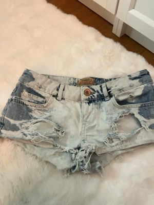 Blå slitna jeansshorts med låg midja - Snygga ljusblå jeansshorts med slitna detaljer och råa kanter. Shortsen har låg midja, klassiska fickor och knäppning fram. Perfekta för en avslappnad sommarstil.