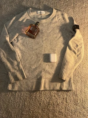 Ljusgrå stickad tröja Stone Island - Ljusgrå stickad tröja från Stone Island Junior med rund halsringning och ribbade muddar. Klassisk patch på ena ärmen. Perfekt för lager-på-lager och enkel att matcha med andra plagg sitter lite töjigt .