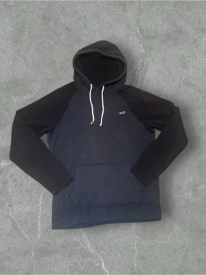 Mörkblå hoodie från Hollister - Snygg mörkblå hoodie från Hollister med svarta ärmar och vit dragsko i huvan. Klassisk känguruficka framtill och Hollisters logga broderad på bröstet. Perfekt för en avslappnad och trendig stil. Tillverkad i mjukt bomullsmaterial.