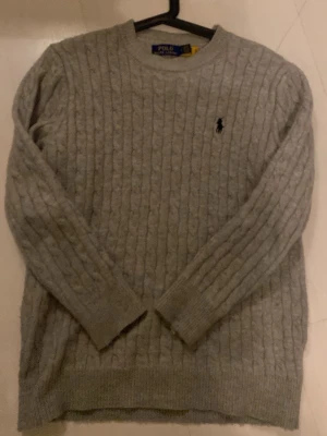 Grå kabelstickad tröja Polo Ralph Lauren - Snygg grå kabelstickad tröja från Polo Ralph Lauren. Tröjan har rund halsringning, långa ärmar och ribbstickade muddar. Perfekt för lager-på-lager och en klassisk look. Materialet är mjukt och håller värmen bra.