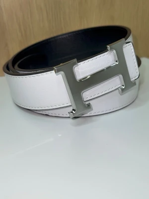 H-Bälte - Snyggt bälte med ett stort, silverfärgat H-spänne. Bältet är 115cm, har vit kontrastsöm längs kanterna och en klassisk, stilren design som lyfter vilken outfit som helst. Bältet är oanvänt (nytt). Du får även med ett armband och ett verktyg för att göra egna hål! Skriv vid minsta lilla fundering!