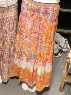 Mönstrad orange långkjol  - Färgstark långkjol från Indiska med paisleyinspirerat mönster i orange, rosa, blått och vitt. Kjolen har volangdetaljer och är tillverkad i ett lätt, luftigt material som passar perfekt för varma dagar. Resår i midjan för bekväm passform. Storleken är onesize men passar xs/s