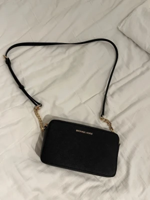 Svart axelväska från Michael Kors - Stilren svart axelväska från Michael Kors med guldfärgade kedjedetaljer och justerbar axelrem. Väskan har en rektangulär form och är tillverkad i ett tåligt material med diskret struktur. Perfekt för dig som vill ha en klassisk och trendig accessoar.