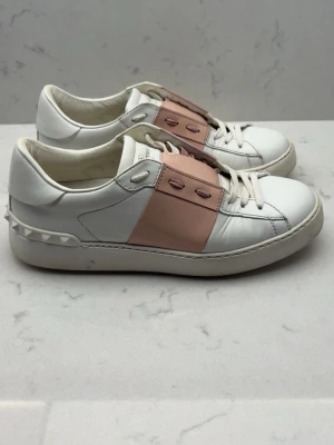 Valentino Garavani sneakers i vitt och rosa - Snygga sneakers från Valentino Garavani i vitt skinn med ljusrosa detalj över snörningen och ikoniska nitar på hälen. Klassisk rund tå och platt sula. Perfekta för dig som vill ha en stilren men ändå unik look. Äkta!!! Behöver säljas snabbt!!!