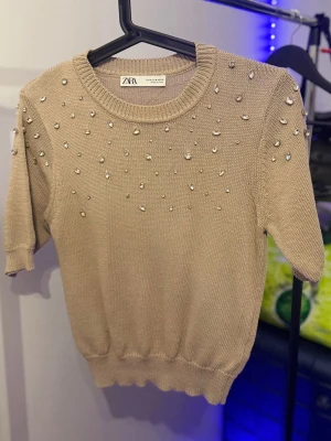 Beige stickad topp med pärlor från Zara - Supersnygg beige stickad topp från Zara med korta ärmar och rund hals. Toppen är dekorerad med glittriga pärlor över axlar och bröst, vilket ger en extra touch. Perfekt att styla med jeans eller kjol för en trendig look.