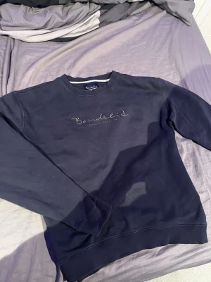 Mörkblå sweatshirt från Bondelid - Mörkblå sweatshirt från Bondelid med broderad logga och texten 'ATHLETIC LEAGUE 56' på bröstet. Tröjan har rund halsringning och ribbade muddar vid ärmslut och nederkant. Perfekt för en avslappnad och stilren look.