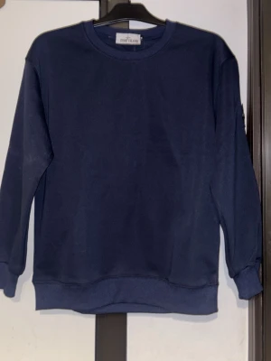 Mörkblå sweatshirt från Stone Island - En stilren mörkblå sweatshirt från Stone Island med klassisk rund hals och ribbade muddar. Tröjan har lång ärm och den ikoniska loggan på vänster ärm. Perfekt för en avslappnad och trendig look. Den är M men är mer som S