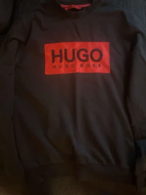 Svart sweatshirt från Hugo Boss - Svart sweatshirt med röd rektangel och svart HUGO-tryck på bröstet. Klassisk rund hals och långa ärmar. Perfekt för en stilren och avslappnad look.