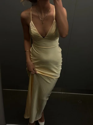 Champagnefärgad långklänning med snörning - Elegant långklänning i champagnefärgad satin med djup v-ringning och tunna axelband. Klänningen har öppen rygg med snygg snörning och figurnära passform. Perfekt för speciella tillfällen när du vill sticka ut.