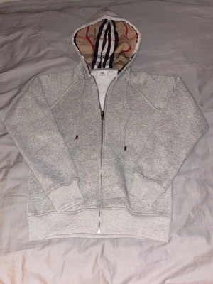 Grå Burberry Zip Up hoodie helt ny  - Grå zip hoodie från Burberry i storlek XS. Klassisk och stilren modell med dragkedja framtill, justerbar huva med Burberrys ikoniska rutmönster på insidan samt diskret broderad logotyp på bröstet. Mjuk och bekväm kvalitet, perfekt för vardagsbruk. Och sprillansny , 