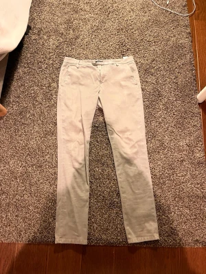 Gråa  chinos från Dondup, stl 33 - Snygga gråa chinos från Dondup i klassisk rak modell. Byxorna har bälteshällor, två sidofickor fram och två bakfickor med knapp. Tillverkade i mjuk bomull med dragkedja i gylfen. Perfekta för en clean och stilren look.