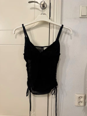 Svart mesh linnetopp med snörning - Svart linnetopp i mesh med smala axelband och snörningsdetaljer på sidorna. Toppen har en lätt v-ringning och är figurnära, perfekt för att skapa en trendig look.