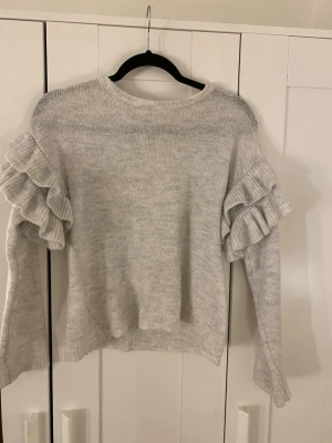 Ljusgrå stickad tröja med volanger - Supersöt ljusgrå stickad tröja med breda volangdetaljer på ärmarna. Tröjan har en avslappnad passform och rund halsringning. Perfekt för dig som vill ha något extra till vardagslooken. Materialet känns mjukt och bekvämt. Knappt använd