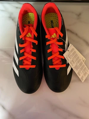 Adidas fotbollsskor Unisex - Snygga Adidas Predator fotbollsskor i svart med vita detaljer och orange snören. Skorna har en modern design med klassiska tre ränder på sidan och greppvänlig sula för bästa fäste på planen. Unisex