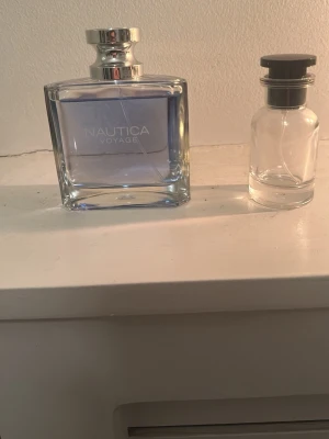 Nautica Voyage parfym blå glasflaska - Fräsch parfym från Nautica, modell Voyage, i en stilren rektangulär glasflaska med silverfärgat lock. Doften har en ljusblå ton och flaskan har en modern, enkel design som känns både klassisk och ungdomlig.