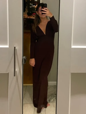 Röd långklänning med öppen rygg - Säljer en elegant brun långklänning med djup v-ringning framtill och öppen rygg. Klänningen har långa ärmar och figurnära passform som ger en stilren siluett. Perfekt för dig som vill sticka ut med en sofistikerad look.