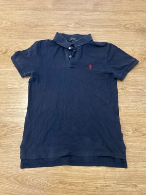 Ralph Lauren Pikétröja - Klassisk marinblå pikétröja från Ralph Lauren med röd broderad logga på bröstet. Tröjan har korta ärmar, krage och knappar framtill. Tillverkad i mjuk bomull för en bekväm känsla och snygg passform.