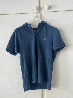 Blå pikétröja från Ralph Lauren - Klassisk blå pikétröja från Ralph Lauren med korta ärmar och vit broderad logga på bröstet. Tröjan har krage och knappar vid halsen. Perfekt till jeans eller shorts för en avslappnad men snygg look.