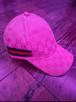 Gucci keps med GG-mönster - Säljer en snygg keps från Gucci i rosa tyg med klassiskt GG-monogram över hela kepsen. På sidan finns en ikonisk röd och marinblå rand. Böjd skärm och justerbar passform. Perfekt för dig som vill ha en lyxig och trendig accessoar.