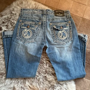 Ljusblå jeans från Live and Fight - Säljer ett par ljusblå jeans från Live & Fight med tydliga kontrastsömmar och broderade bakfickor. Jeansen har coola detaljer som knappar och dekorativa nitar. 