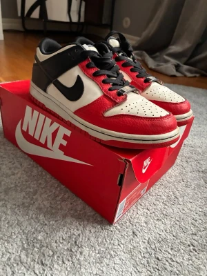 Nike Dunk Low röd/svart/vit - Nike Dunk Low sneakers i rött, svart och vitt skinn. Klassisk låg modell med svart swoosh, snörning och vit mellansula. Snygg kontrast mellan färgerna och retrovibe. Perfekta för dig som vill sticka ut med dina sneakers. Nypris 2400kr