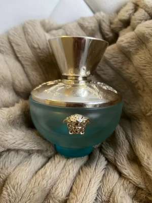 Versace turkos parfymflaska - Säljer en lyxig parfym från Versace i en rund, frostad turkos flaska med silverfärgat lock och detaljer. Flaskan har Versaces ikoniska Medusa-huvud i silver på framsidan och ett elegant mönster på locket. Perfekt för dig som gillar exklusiva dofter.