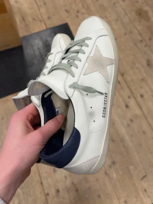 Golden Goose vita sneakers med stjärna - Snygga vita sneakers från Golden Goose med grå mockastjärna på sidan, grå snörning och mörkblå häl. Skorna har en klassisk rund tå och platt sula. Perfekta för dig som gillar en trendig och avslappnad stil. Skick 10/10 nästan helt nya. Box osv tillkommer. Även certifikat. Hör av vid frågor!😊