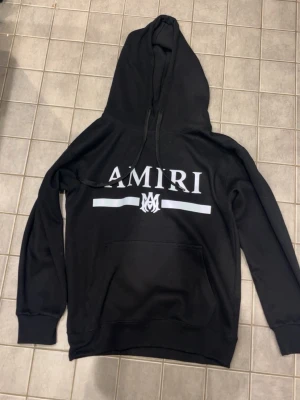 Svart hoodie från Amiri med tryck - Svart hoodie från Amiri med stor vit logga och grafiskt tryck på bröstet. Klassisk känguruficka framtill, dragsko i huvan och ribbade muddar. Perfekt för en streetwear-look och riktigt skön att bära.