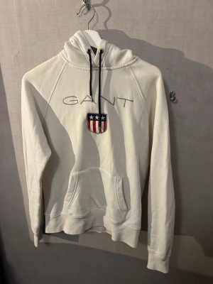 Vit hoodie från GANT med logga - Snygg vit hoodie från GANT med broderad logga och amerikanskinspirerat sköldmärke på bröstet. Hoodien har huva med svarta dragsnören och en stor magficka framtill. Tillverkad i mjukt material och har ribbade muddar vid ärmar och nederkant.