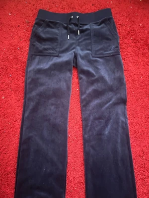 Juicy Couture Mörkblå mjukisbyxor  - Mörka svarta mjukisbyxor i mjuk sammet med bred resår i midjan och snörning framtill. Stora fickor fram och bakfickor med knappdetaljer ger en chill och avslappnad vibe. Perfekta för att maxa myset hemma eller när du vill vara bekväm.