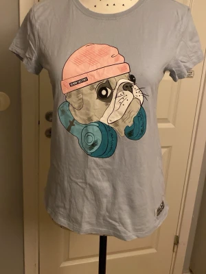 Ljusblå t-shirt med bulldog-tryck - Säljer en ljusblå t-shirt med ett stort tryck av en bulldog som har rosa mössa och blå hörlurar. T-shirten har rund halsringning och korta ärmar. Perfekt för dig som gillar coola och unika motiv!