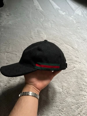 Svart keps med GG-mönster - Snygg svart keps med klassiskt GG-mönster i tyget och en ikonisk grön-röd rand på sidan. Justerbar rem bak för perfekt passform. Perfekt accessoar för dig som vill ha en exklusiv och trendig look.