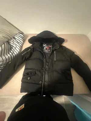 Svart dunjacka med huva från Moose Knuckles - Säljer en svart dunjacka från Moose Knuckles med huva och pälskant. Jackan har flera dragkedjor, fickor med knapp och dragkedja samt quiltad design. Perfekt för kalla vinterdagar och har en snygg, sportig look. BYTE OCKSÅ 