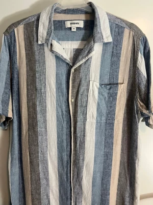 Randig skjorta från Kappahl i linne - Säljer en snygg randig skjorta från Kappahl i relaxed fit. Skjortan har vertikala ränder i blått, beige, grått och vitt, korta ärmar, bröstficka och knappar framtill. Perfekt för en chill och stilren look.