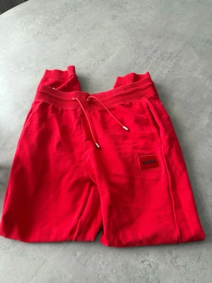 Röda mjukisbyxor från HUGO - Snygga röda mjukisbyxor från HUGO med ribbad midja och snörning. Byxorna har en liten HUGO-logga på benet och är tillverkade i mjukt bomullsmaterial. Perfekta för en chill och avslappnad stil.