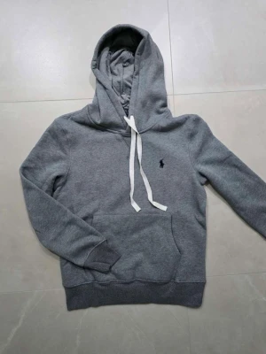 Grå hoodie från Polo Ralph Lauren - Snygg grå hoodie från Polo Ralph Lauren med klassisk huva och vita dragsnören. Tröjan har en känguruficka framtill och den ikoniska lilla loggan broderad på bröstet. Tillverkad i mjukt bomullsmaterial som är perfekt för chill dagar.